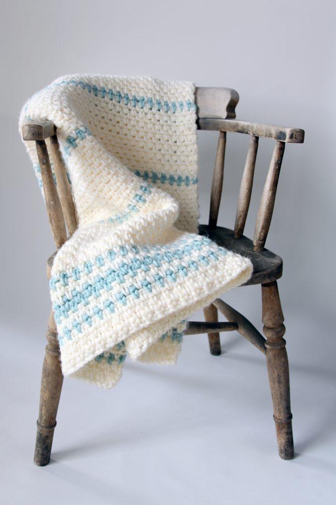 Moss stitch crochet blanket on a wooden chair in cream and blue yarn.