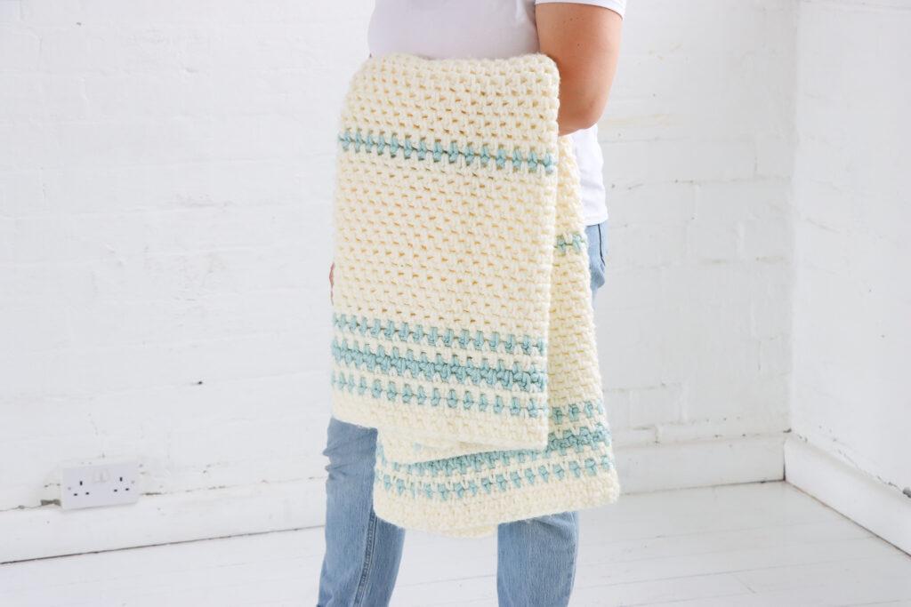 female stood in a white room with a crochet blanketing colours cream with a pale blue stripe draped over her arm. 