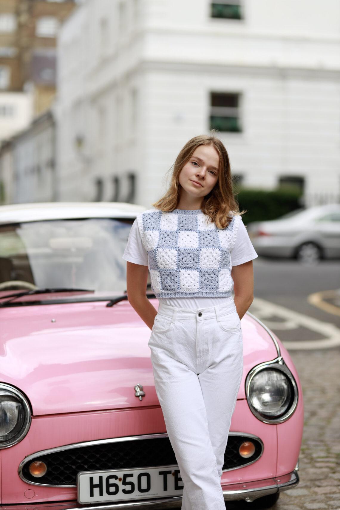 a female stood in front of a pink car in the street wearing white jeans and a crochet granny square t.shirt in white and blue granny squares.