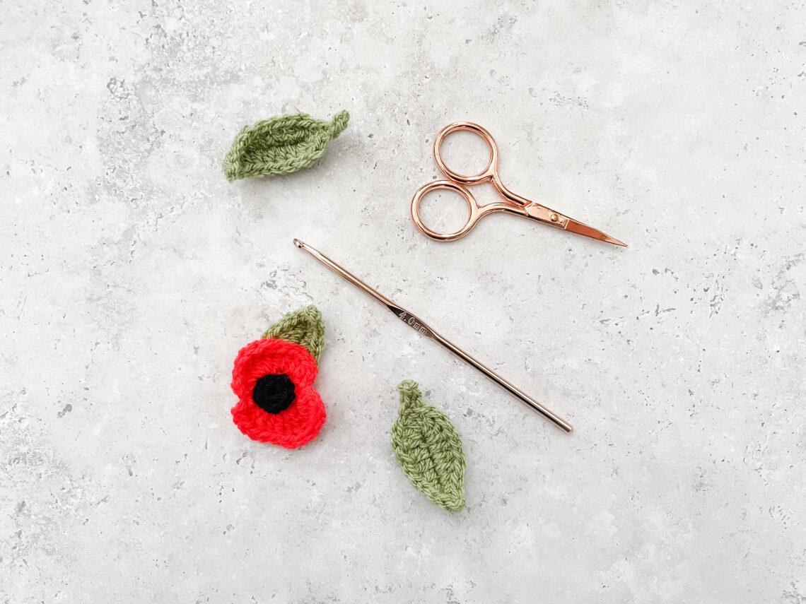 a flat lay image of a crochet poppy lay alongside two crochet leaves a rose gold hood and scissors lay on a concrete effect back drop.