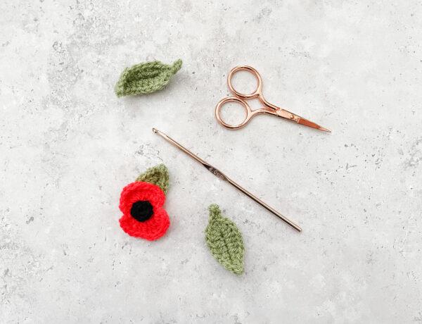a flat lay image of a crochet poppy lay alongside two crochet leaves a rose gold hood and scissors lay on a concrete effect back drop.