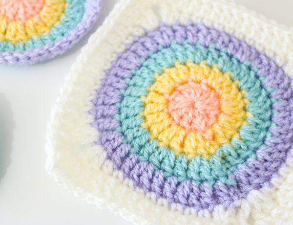 Flat lay image of a circular granny square. The circle is made up of four different colours: peach, yellow, teal and purple with a cream border squaring off the circular shape.