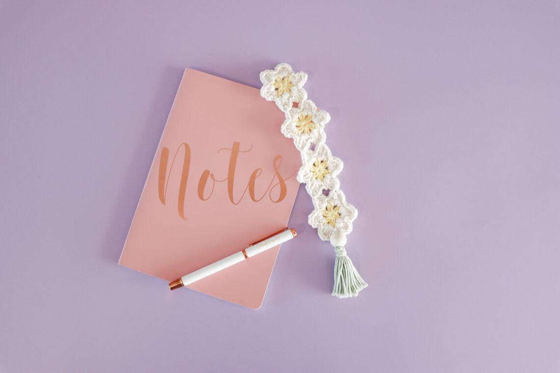 A daisy crochet bookmark in a chain with a tassel on the end in yellow, white and green yarn pictured beside a pink notebook with 'notes' on the front and a white pen on top.