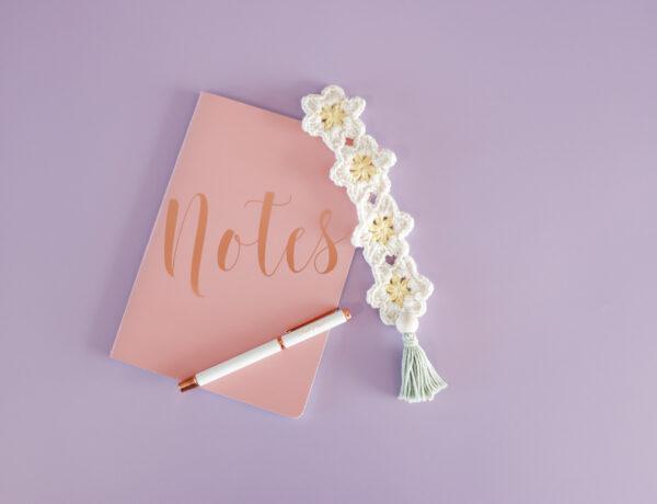 A daisy crochet bookmark in a chain with a tassel on the end in yellow, white and green yarn pictured beside a pink notebook with 'notes' on the front and a white pen on top.