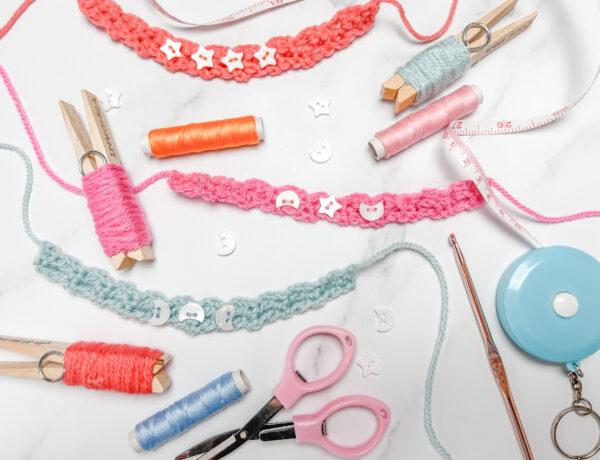 flay lay image on a marbled backdrop showing three crochet friendship bracelets in pink, orange and pale blue, yarn pegs, embroidery thread, scissors, crochet hook and tape measure.