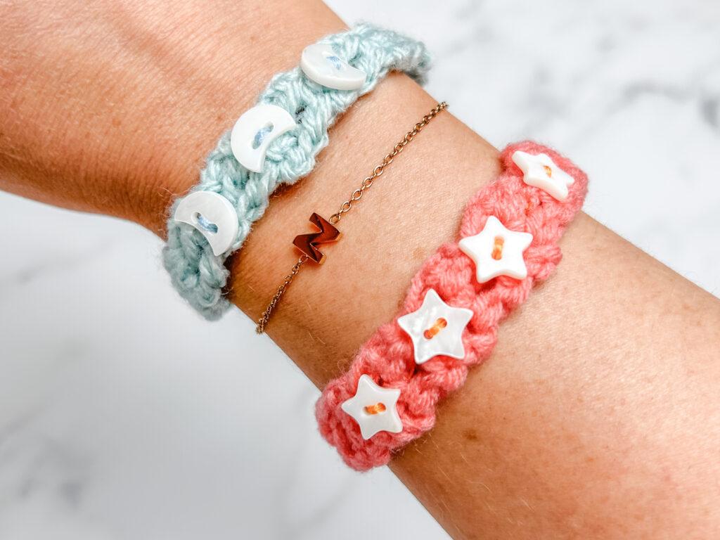 two crochet bracelets modelled on a wrist with a white marbled background. One orange with star beads attached and the other blue with white moon beads attached. A rose gold bracelet sits in the middle with the initial N. 