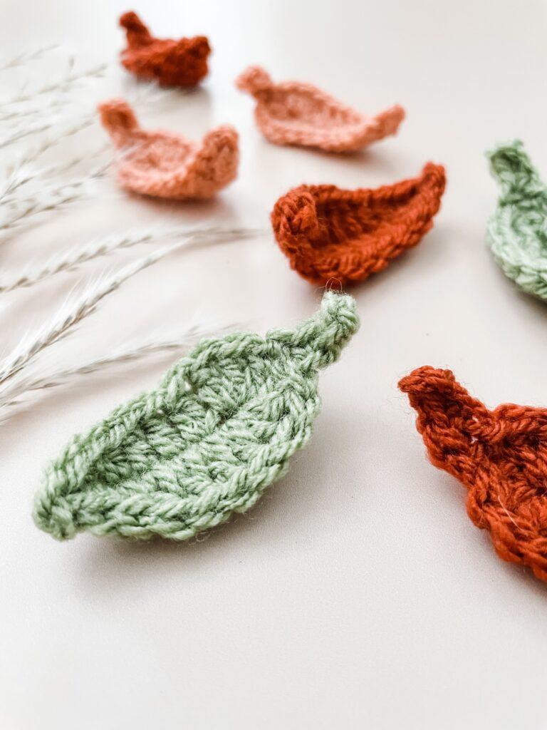 Crochet leaves on a nude background in green, rust and peach yarn with pampers grass appearing in the background. 