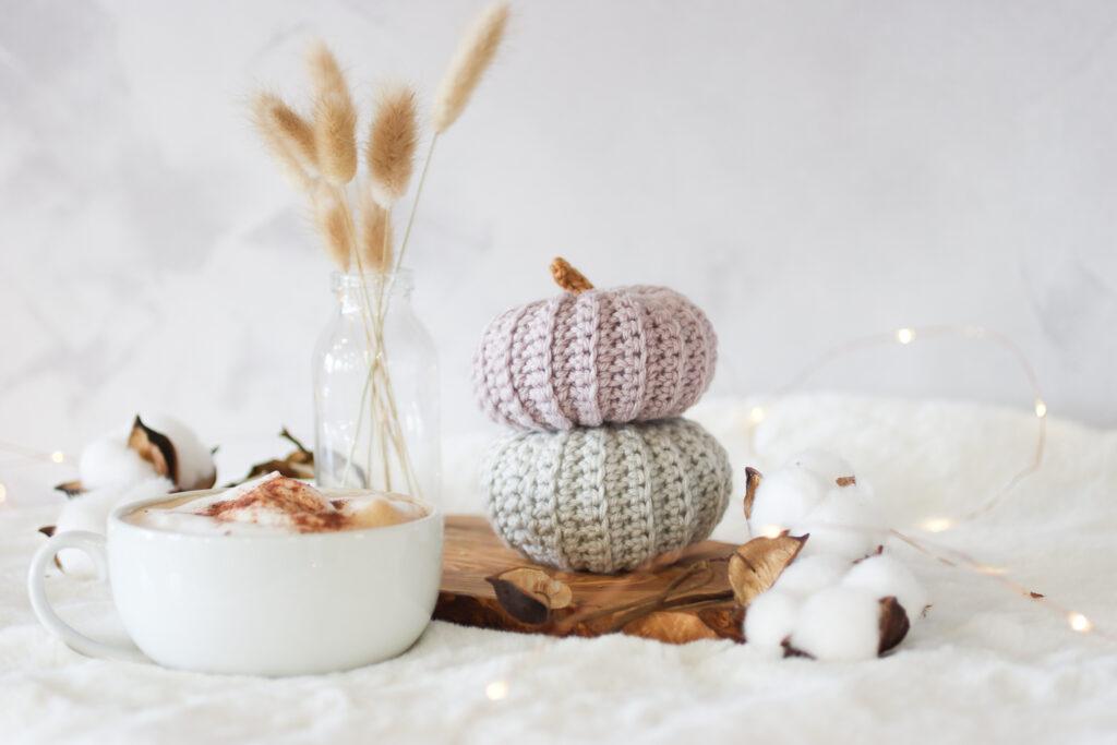 two crochet pumpkins stacked one grey and the other in mushroom. The crochet pumpkins are surrounded by small fairy lights with a cup of coffee and bunny tail dried flowers in a small glass vase. the background  is white and the set up is placed on a white faux fur rug