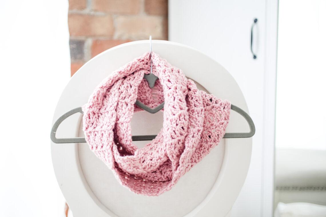 a pink infinity scarf wrapped around a grey coat hanger hooked on the back of a white chair