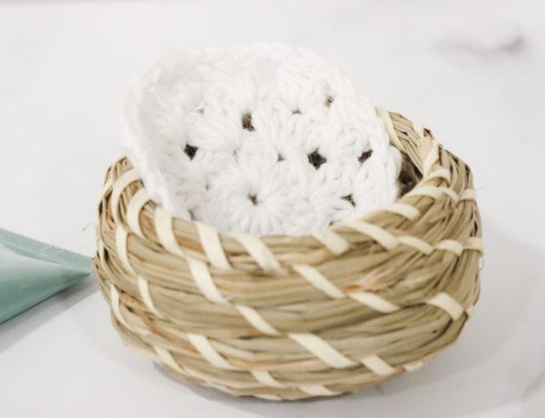 crochet face scrubbie made from white cotton yarn sat in a small wicker basket