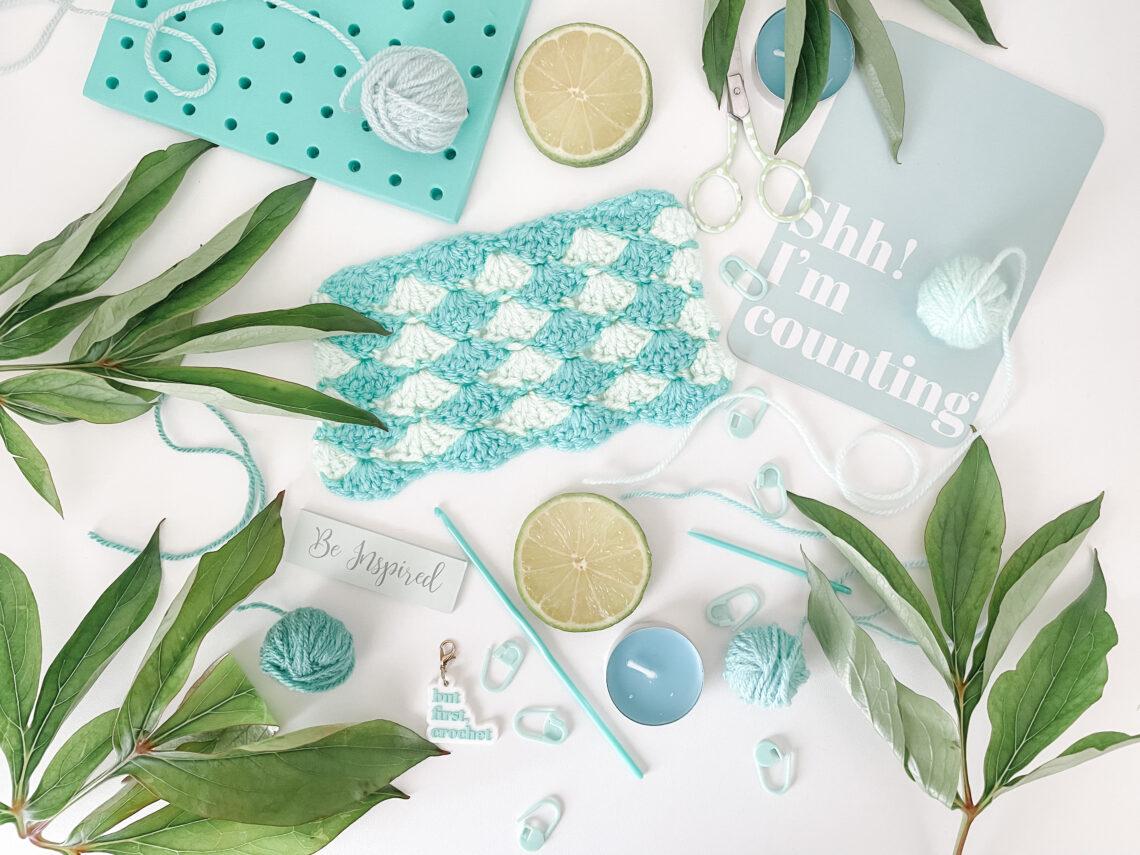 A blue crochet swatch surrounded by a blocking board, candles and plants in a flat lay style on a white background.