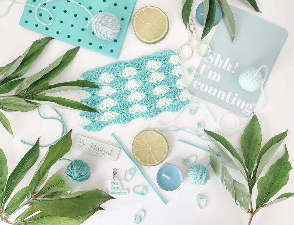 A blue crochet swatch surrounded by a blocking board, candles and plants in a flat lay style on a white background.