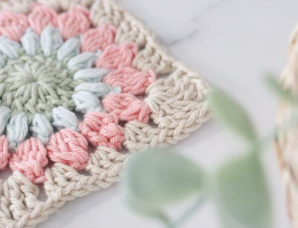 a sunburst granny square lay on a marble backdrop at a slight angle with foliage in the bottom right side corner of the image. The sunburst granny is made from pale green, blue, pink and beige.