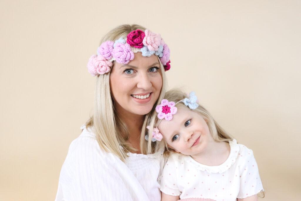 an image of mother and daughter sat infront of a cream backdrop wearing a summer crochet flower crown each made in pink, purple and pale blue yarn.