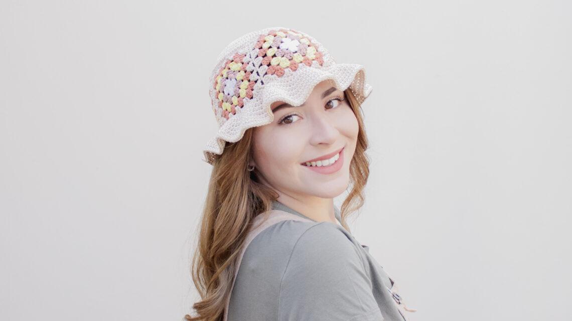 an image of a crochet bucket hat with a frilled rim made up of granny squares worn by a female. The hat is cream and the granny squares are made up of shades of browns into yellow.