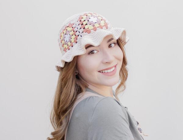 an image of a crochet bucket hat with a frilled rim made up of granny squares worn by a female. The hat is cream and the granny squares are made up of shades of browns into yellow.