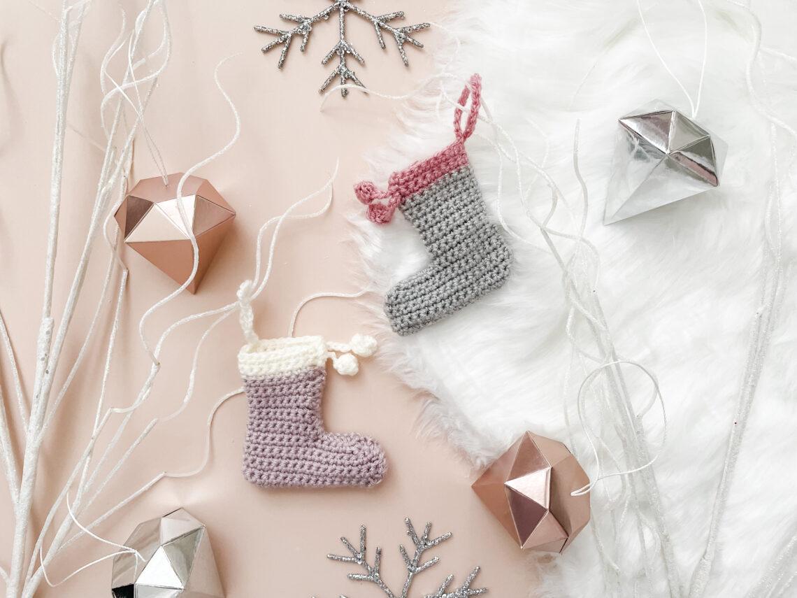 two mini crochet stockings lay flat on a pale pink backdrop with a fur rug to the right hand side of the image surrounded by white sparkly branches, snowflake and diamond-shaped decorations