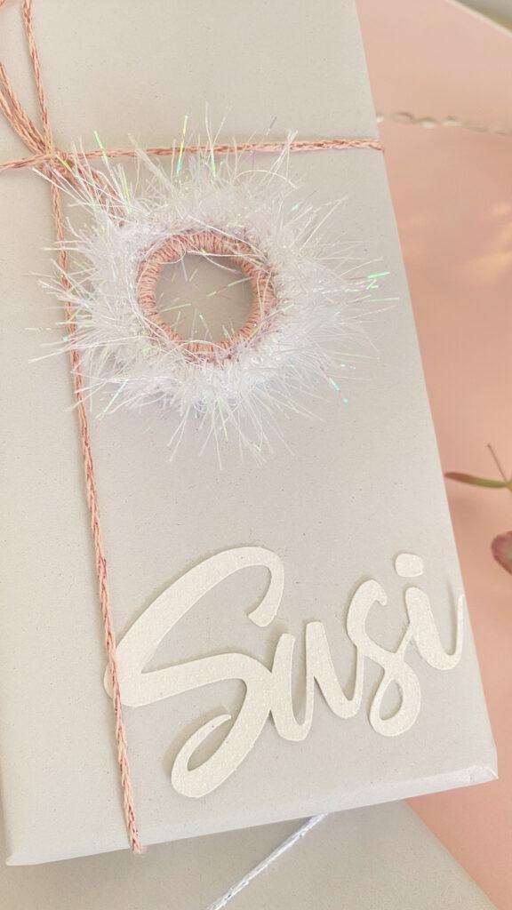 sparkly crochet present topper pictured attached to a wrapped gift lay flat on a pale pink background.