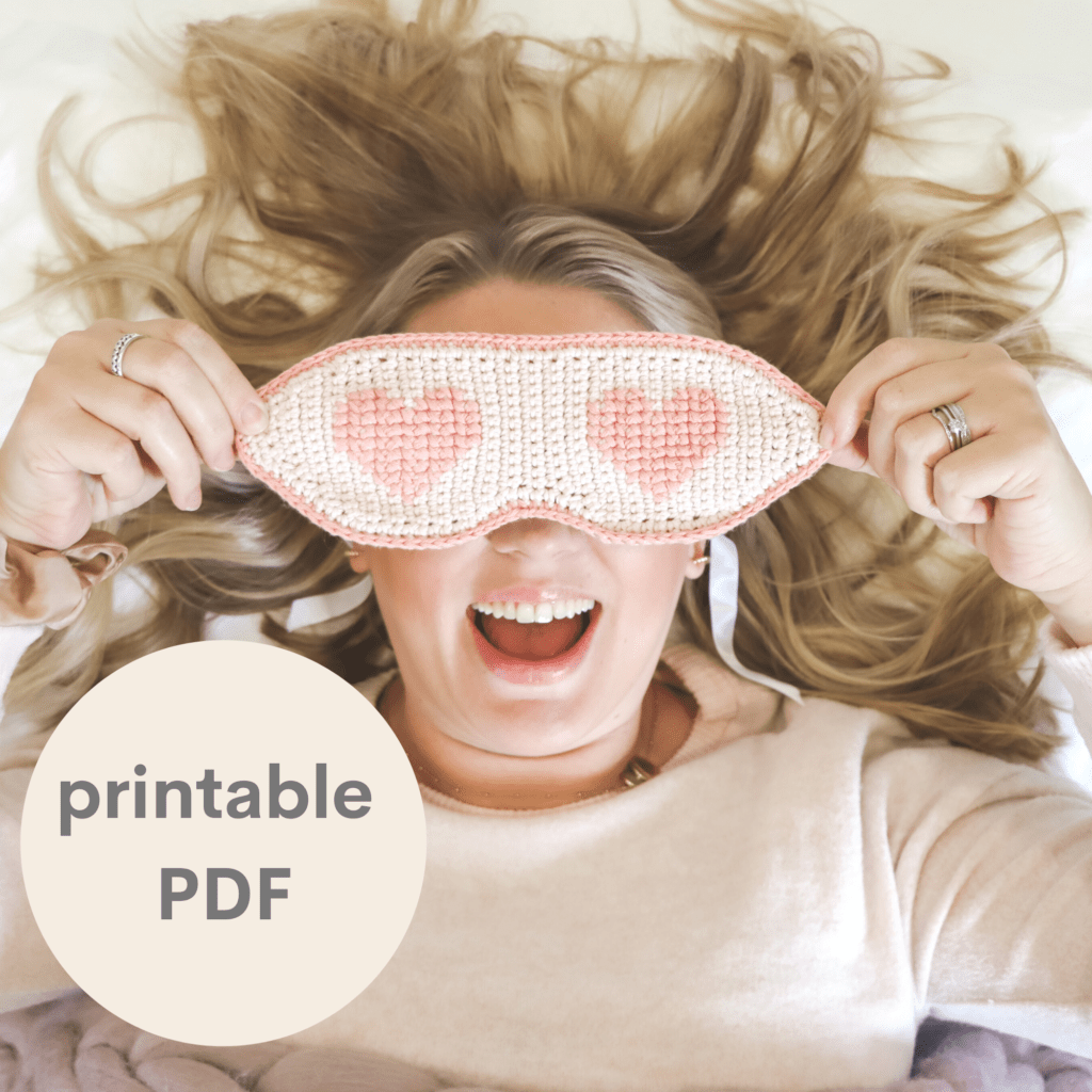 A lady laid down wearing a crochet eye mask with hearts on the eyes.