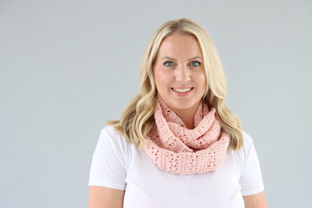 female stood in front of a grey background wearing a white t.shirt and a pale pink crochet infinity scarf.