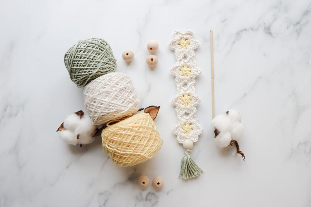 A Crochet Daisy Bookmark laid on a marble background alongside a wooden crochet hook and 3 yarn cakes in green, white and yellow.