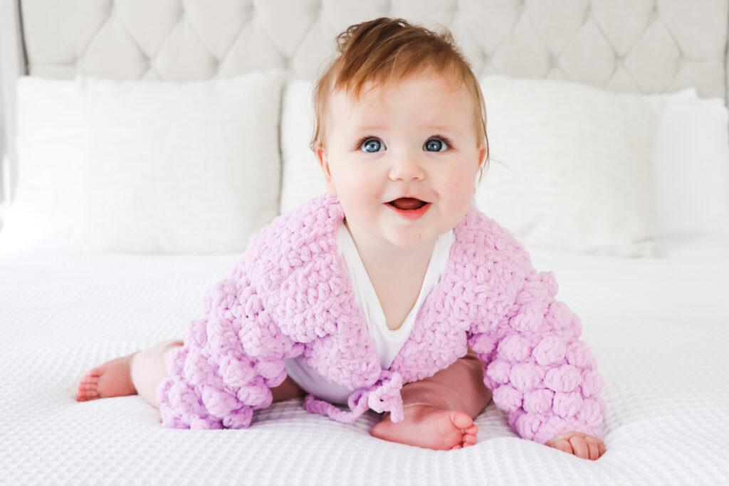 baby girl sat on a white bed wearing a textured baby bolero in lilac yarn. 