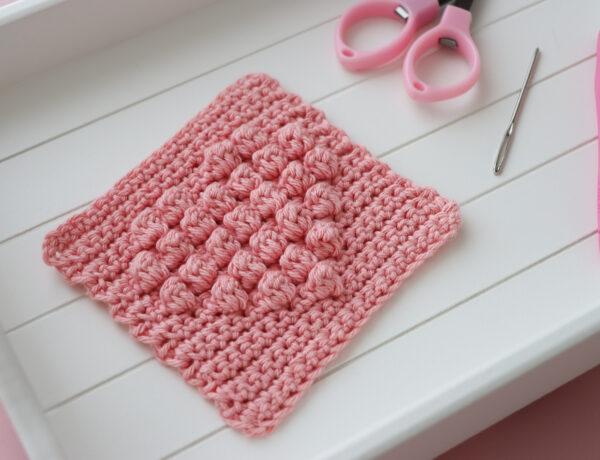 a crochet bobble heart square on a white wooden tray alongside crafting scissors and a darning needle.