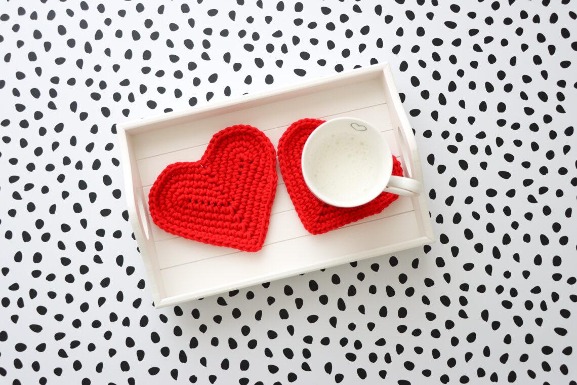Two red crochet heart coasters on a white wooden tray. One with a mug on.