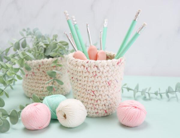 crochet nesting baskets on a desk one with a hanging plant sat inside and the other filled with pink crochet hooks and pastel green pencils. small balls of yarn are sat infant of the nesting baskets on the desk.