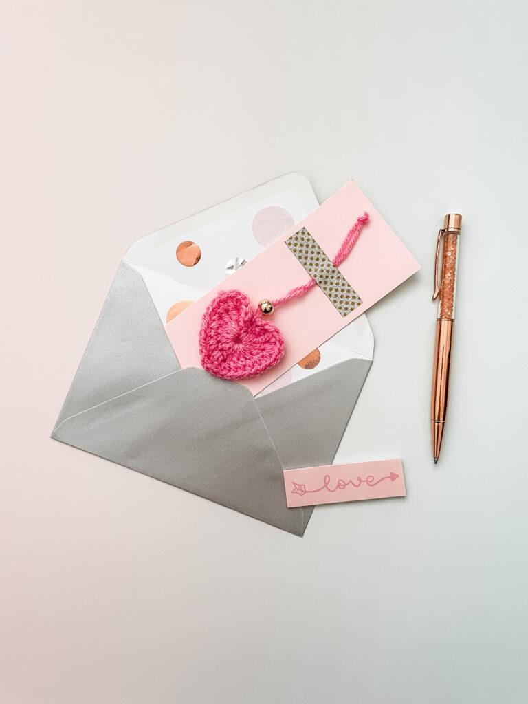 A crochet heart falling out of an envelope with a pen next to it as a heart charm with love note.