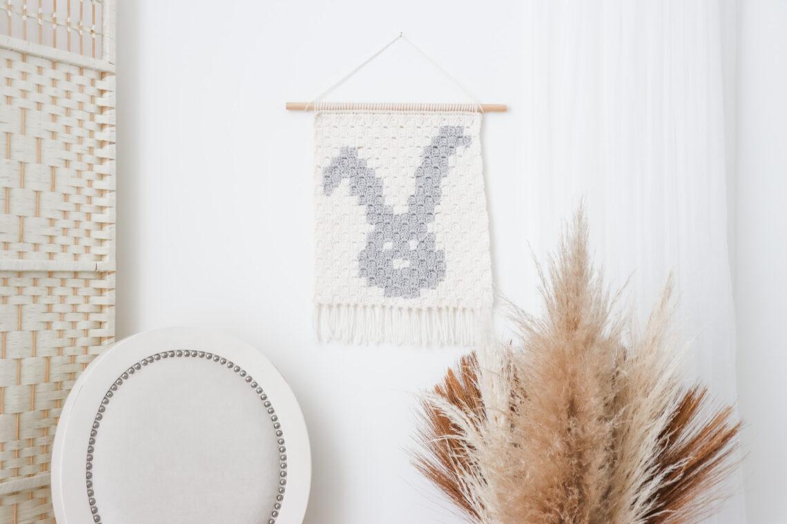 A bunny wall hanging using the corner to corner crochet method on a white wall with plants and a chair surrounding it.