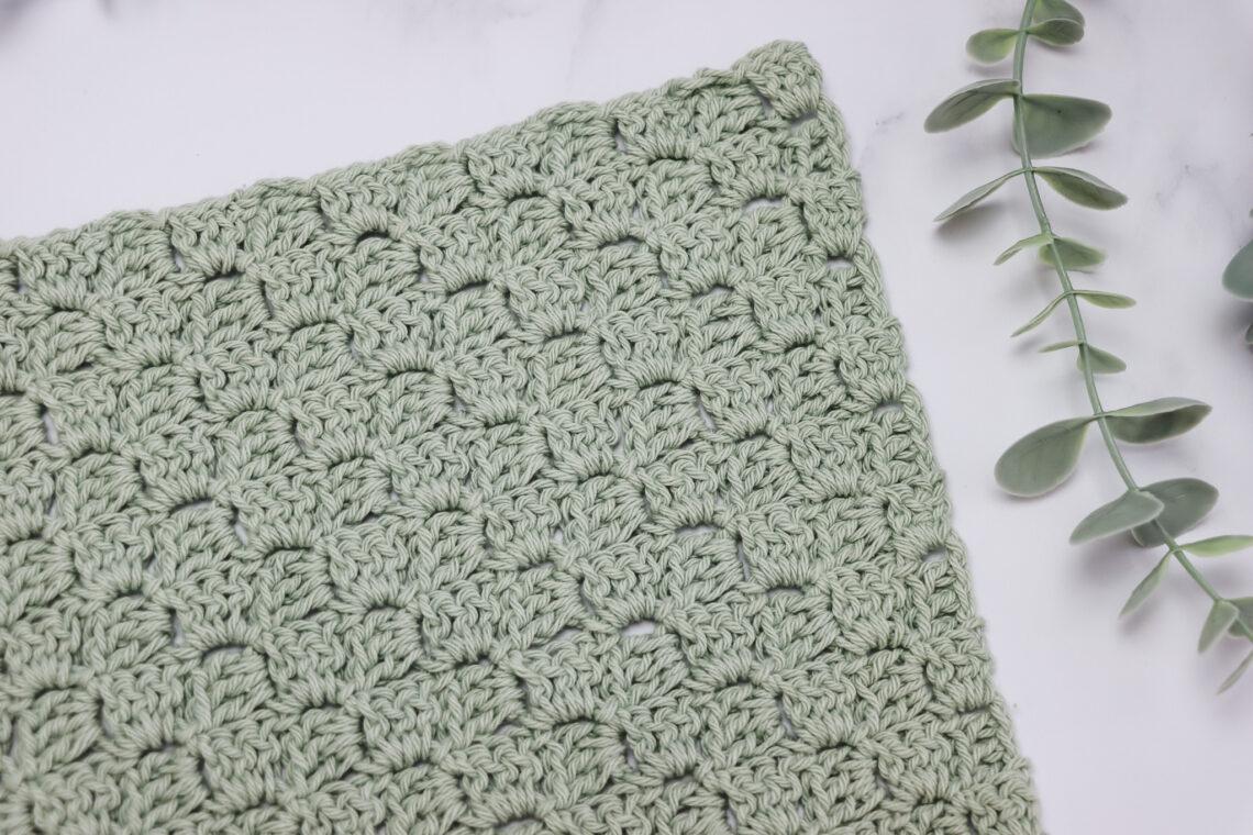 crochet swatch of the corner to corner stitch in a pale green yarn lay flat on a white marble backdrop with foliage in the top right corner of the image.