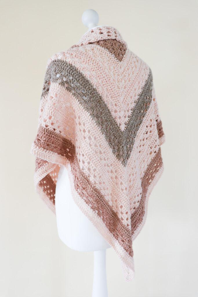 a crochet shawl made is neutrals shades of metallic yarn worn on a white mannequin stood in front of a cream background