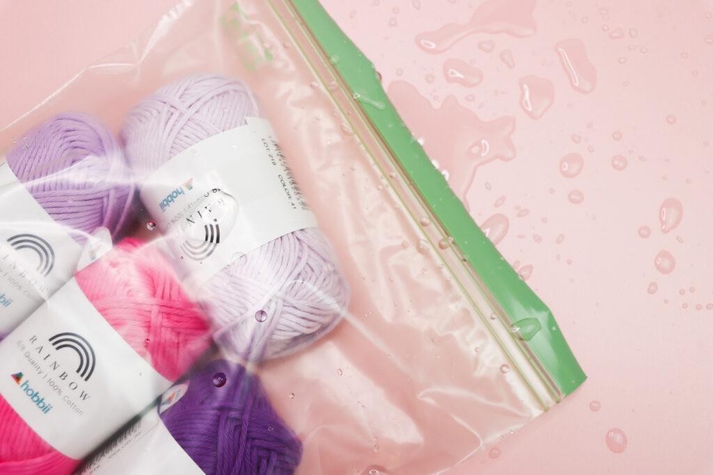 yarn inside a plastic sealed bag lay flat on a pink background with water splashed around it to show the yarn being kept dry by the pool when crocheting on holiday. 