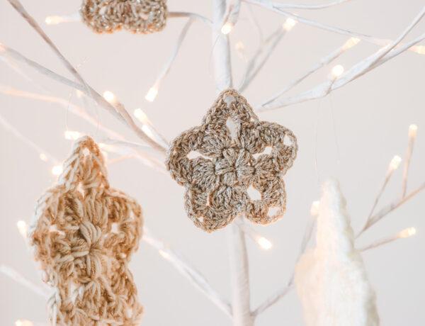 four crochet stars made in gold, cream and neutral sparkly yarn hanging from a white branch tree that has lights on the tips of the branches