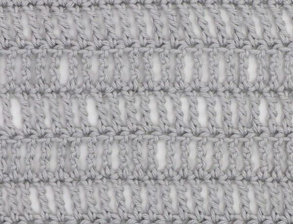 Close-up of a crochet fabric made using the triple treble stitch, showing tall, elongated stitches in light gray yarn with visible vertical gaps for an airy, lacy texture.